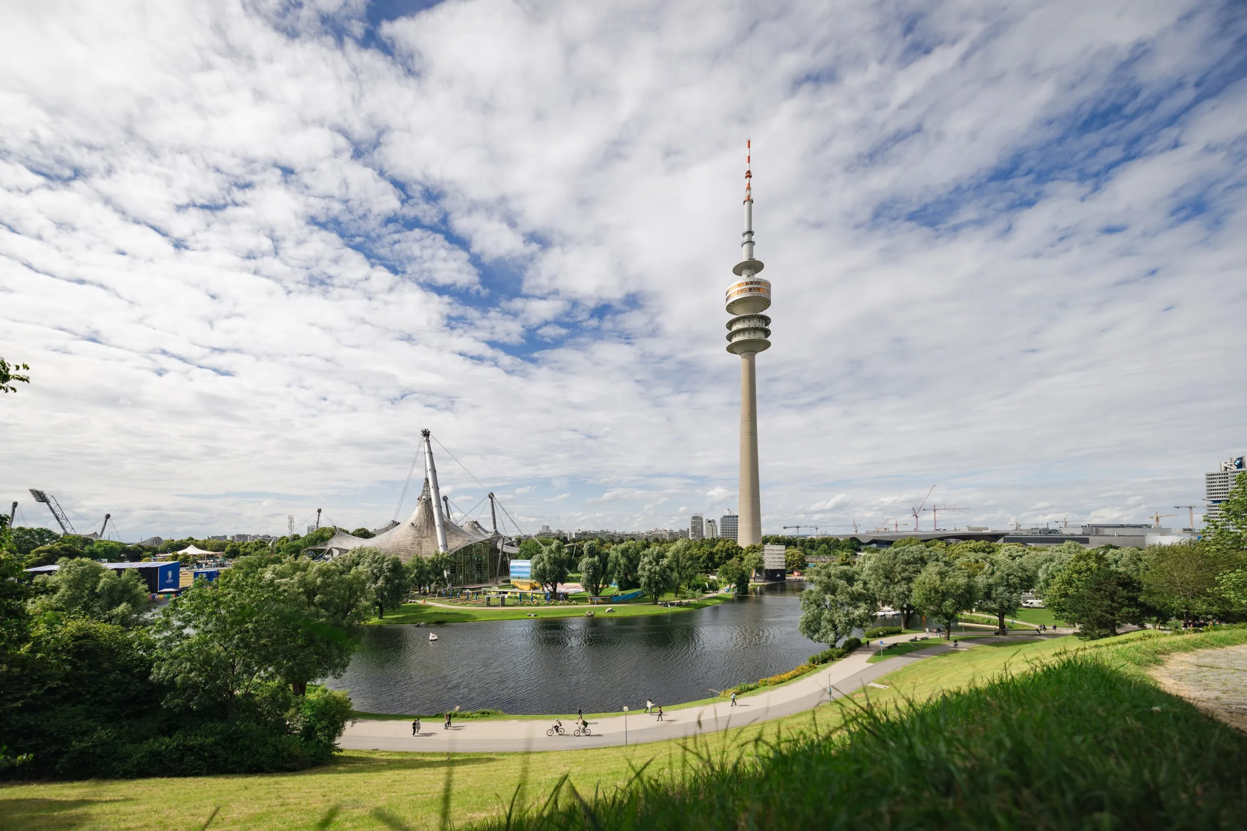 A TV tower rising over a park lake under a sky full of broken white cloud