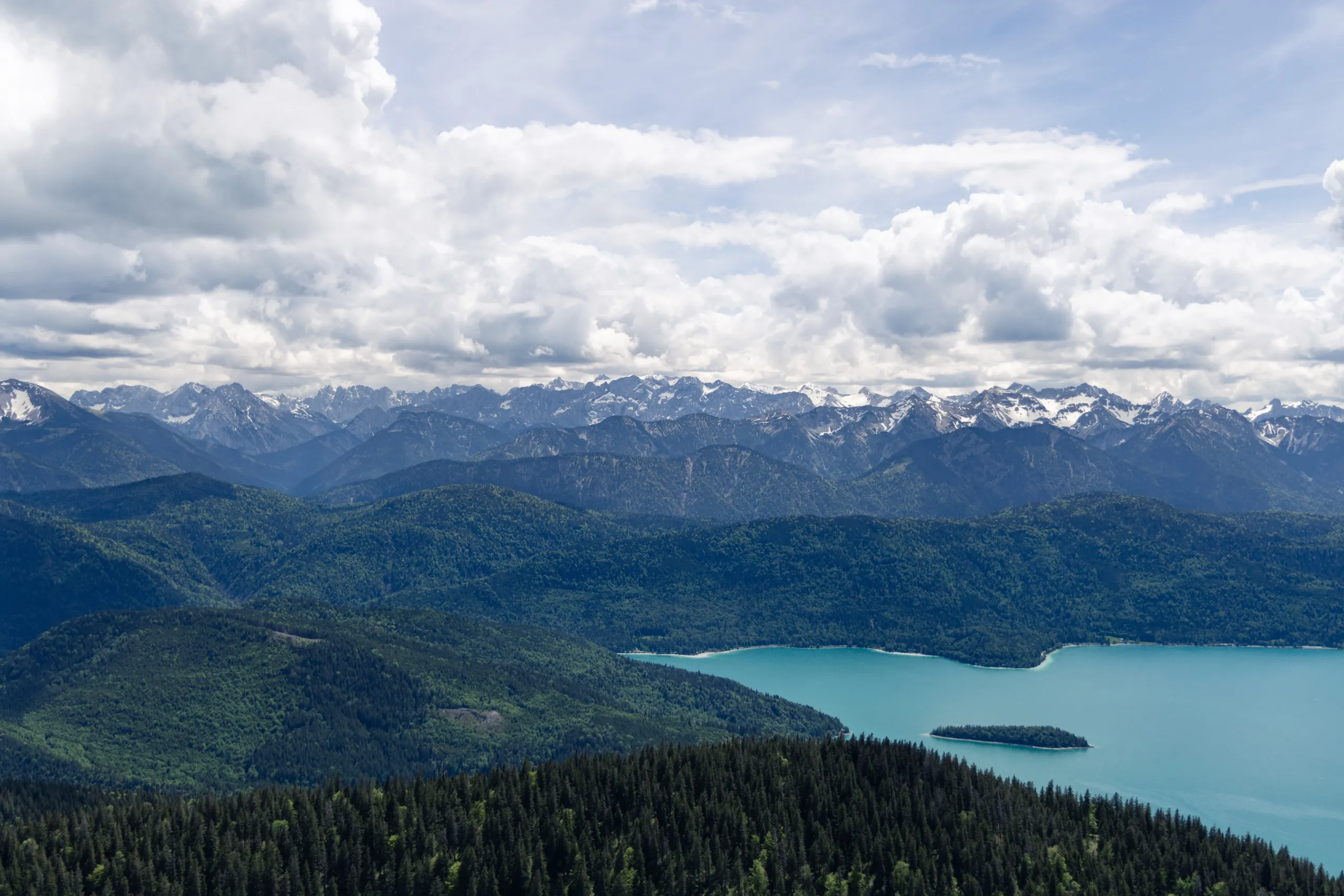 A turquoise alpine lake below forested hills and snow-capped mountains