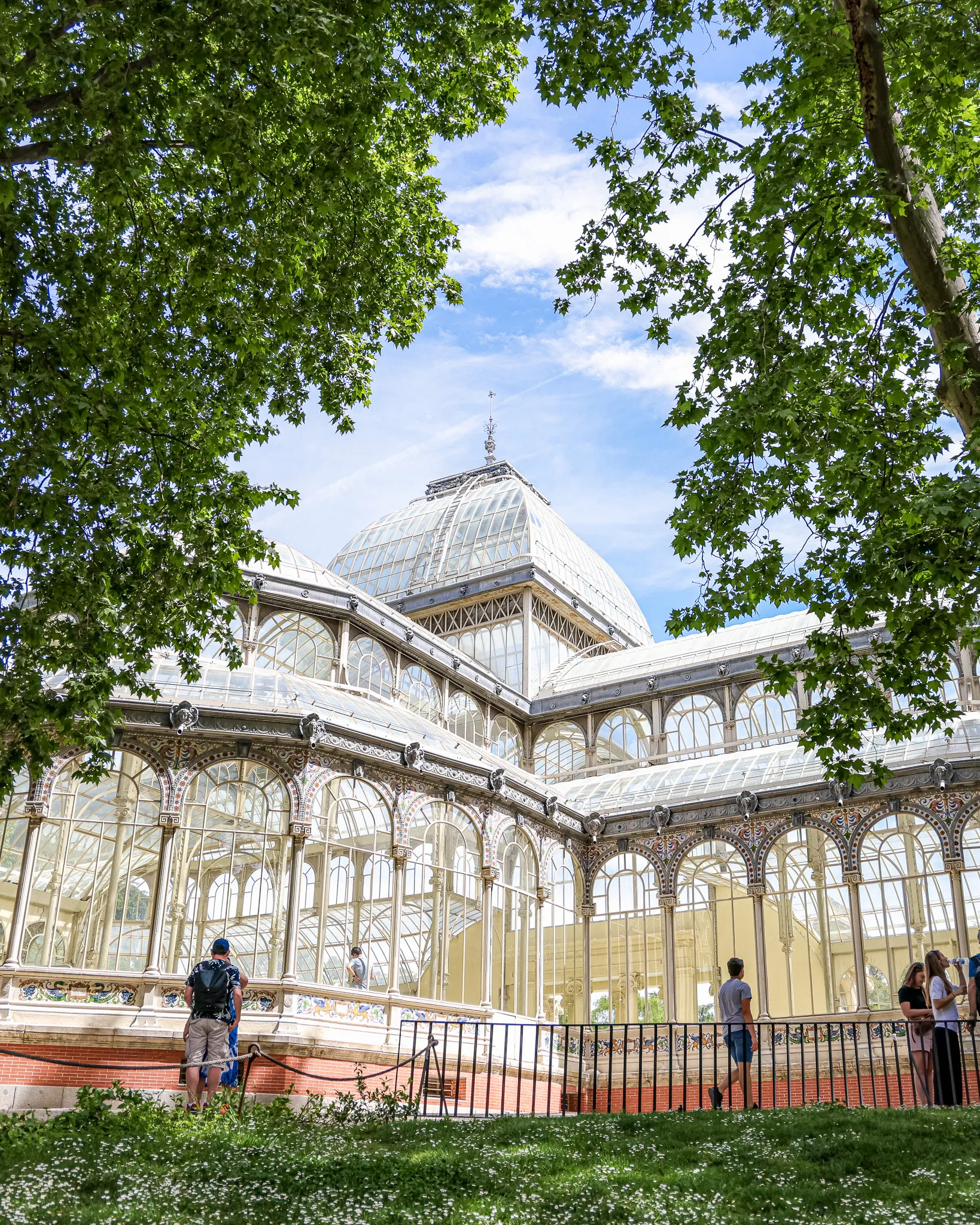 A glass and iron palace framed by green branches, visitors standing in front of it